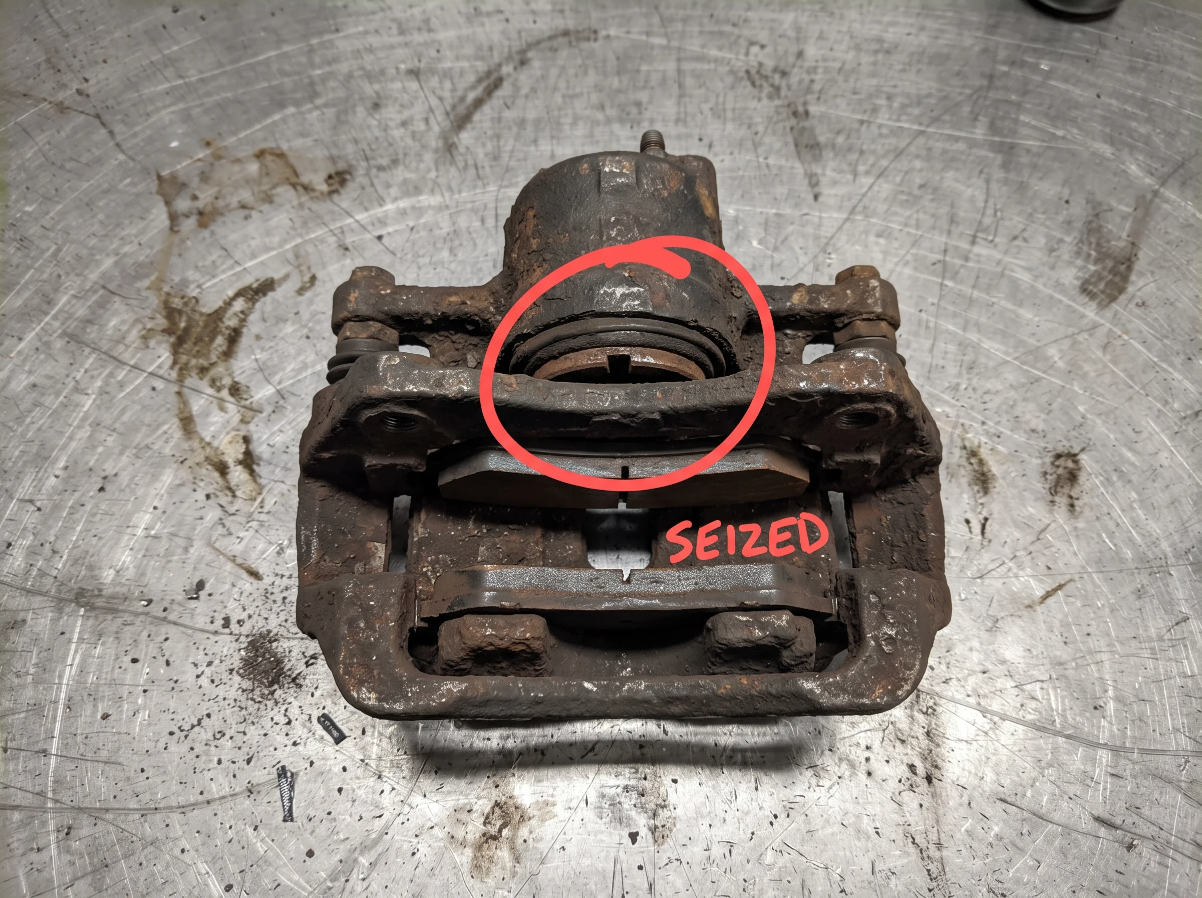 A corroded rear brake caliper on the bench with the seized slider circled in red marker